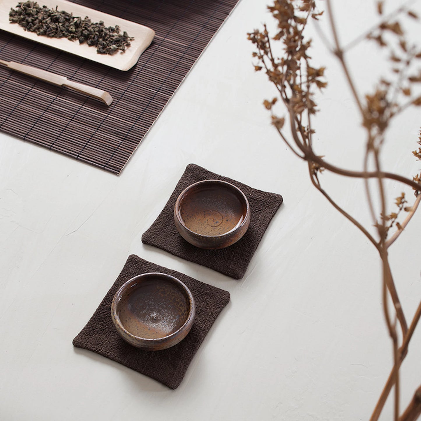 Top-down view of two dark brown fabric tea mats with ceramic cups, arranged on a minimalist white surface with tea accessories