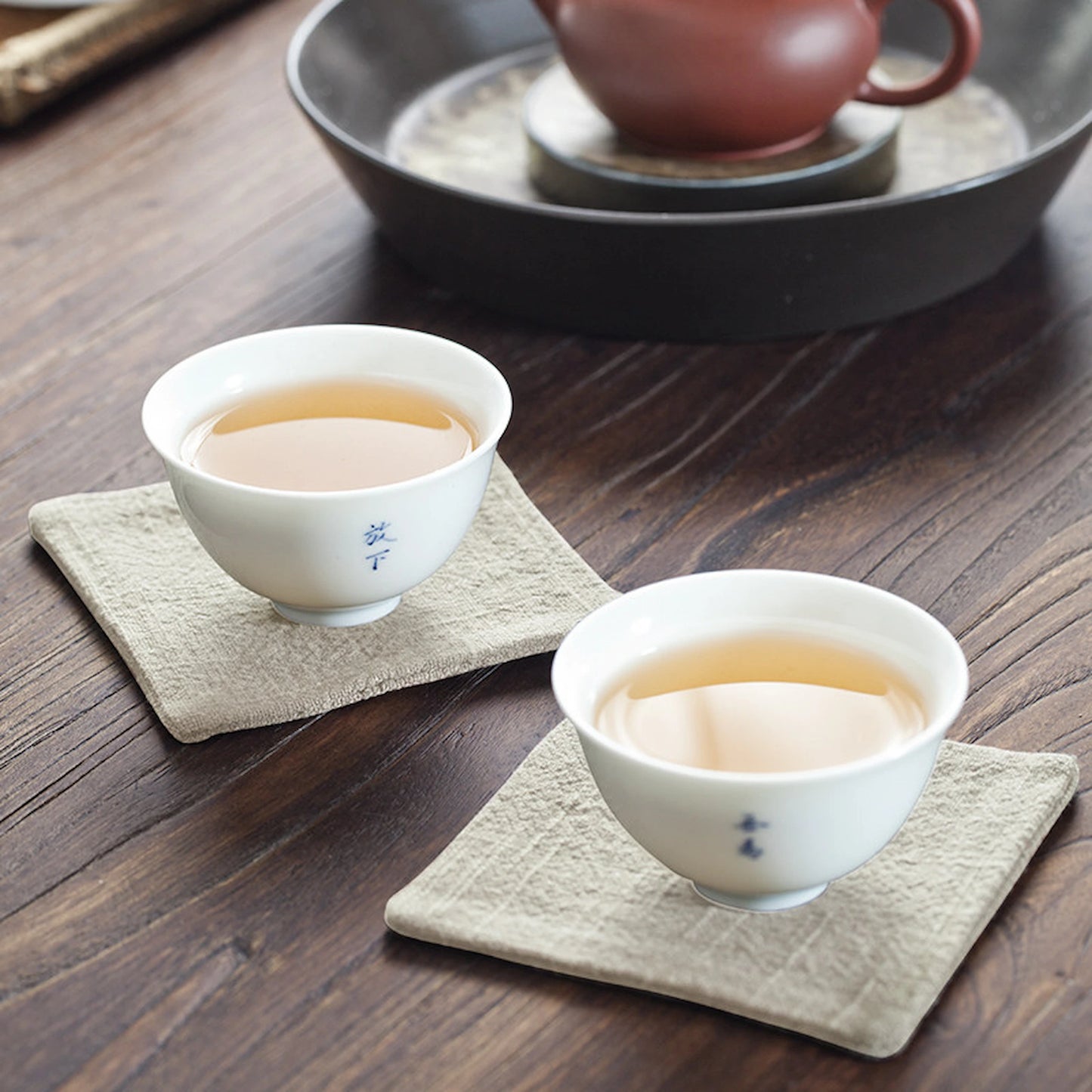Two light beige linen coasters holding white porcelain tea cups on a dark wood table, creating a serene Japanese tea ceremony vibe