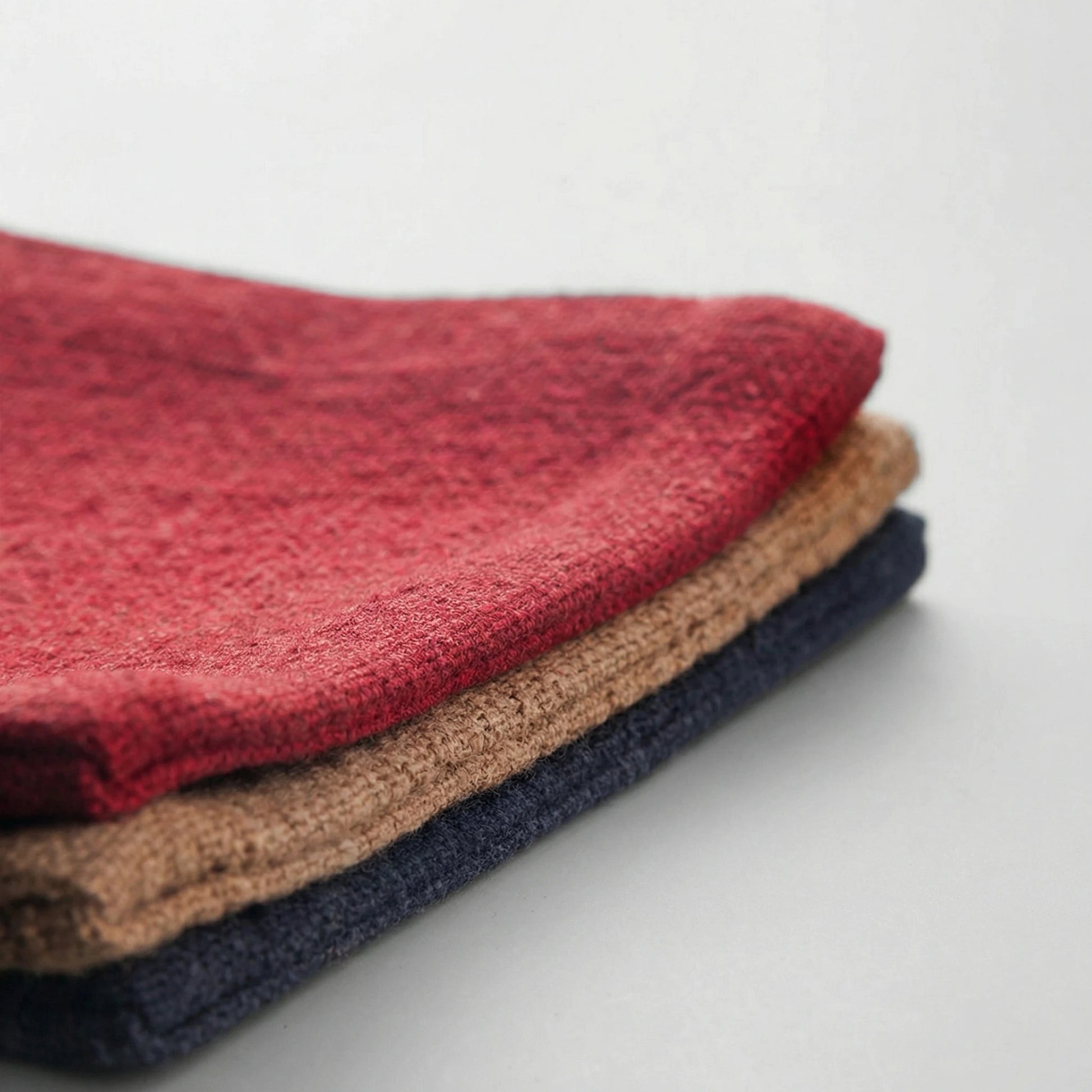 Detail shot of three folded fabric tea mats in red, tan, and navy blue, showing the soft, durable hand-sewn edges